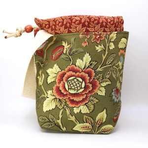 May include: A fabric drawstring pouch featuring an olive green base with a large floral pattern in shades of red, orange, and cream. The interior lining is a contrasting rust-colored fabric with a small leaf print, and a beige cord with a bead.