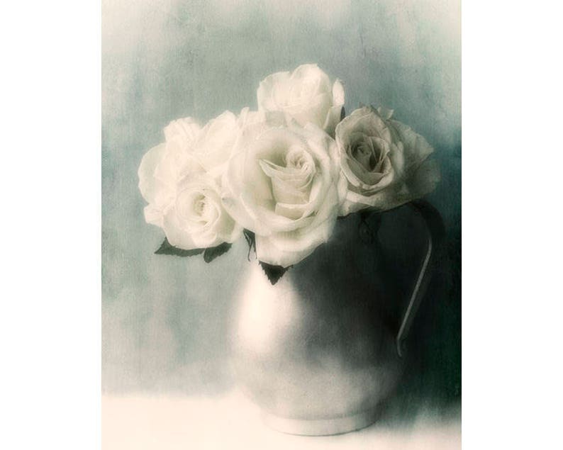May include: A bouquet of white roses in a vintage, silver pitcher. The roses are arranged in a soft, romantic style, with their petals gently overlapping. The background is a soft, muted blue.