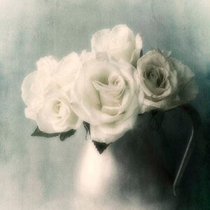 May include: A bouquet of white roses in a white ceramic pitcher. The roses are arranged in a cluster, with the stems extending out of the pitcher. The background is a soft blue.