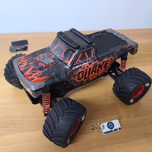 May include: A black and red remote control monster truck toy with the word "QUAKE" on the side. The truck has large black tires with red accents and orange suspension springs. A small white tag with text is next to the truck.