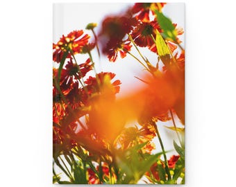 May include: A hardcover journal with a white cover, featuring a vibrant image of red and orange flowers with green leaves. The flowers are in sharp focus, with a bright, sunny background. The journal is a perfect gift for writers and artists.