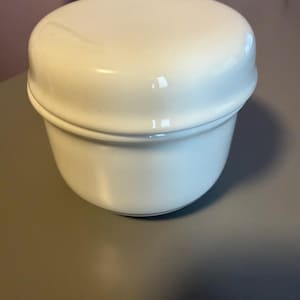 May include: A glossy white ceramic jar with a matching rounded lid. The container features a smooth, cylindrical body with a slightly tapered base and a distinct horizontal ridge where the lid meets the main vessel, creating a clean, uniform appearance.