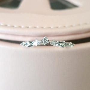 May include: A delicate silver ring featuring a series of marquise-cut gemstones and a central, pointed diamond. The ring's design is elegant and minimalist, perfect for everyday wear or special occasions. The ring is set against a soft pink background.