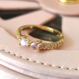 May include: A gold-colored ring with a bow design featuring two heart-shaped, light pink gemstones. The ring band is encrusted with small, clear gemstones. The ring is set against a soft pink background.