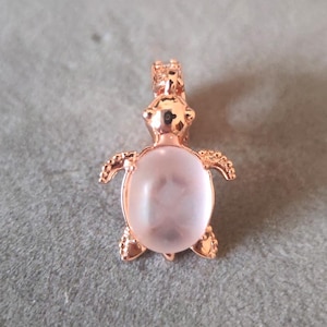 Op de afbeelding: Een roze gouden schildpad hanger met een witte opaal steen in het midden. De schildpad is versierd met kleine accenten rond de randen.