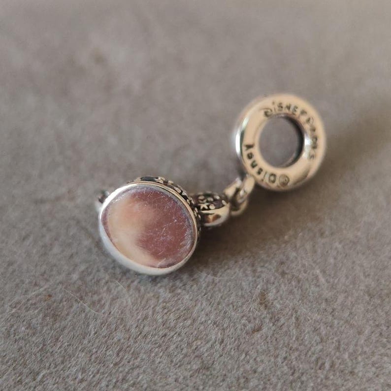 May include: A silver charm with a round, light pink center and intricate detailing. The charm has a loop with the word "Disney" engraved on it. The charm is likely a jewelry accessory.