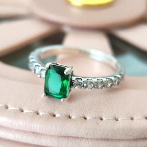 May include: A silver ring with a large, emerald-cut green gemstone as the focal point. The band is adorned with smaller, clear gemstones. The ring is set against a soft pink background, highlighting the jewelry's sparkle and design.
