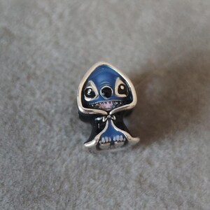 May include: A silver charm featuring a blue alien character with large eyes and a mischievous grin. The charm has a dark blue head and body, with a silver outline. The charm is likely a collectible or accessory.