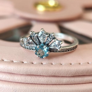 May include: A silver ring with a cluster of clear and blue gemstones. The ring features a central, round, light blue gemstone surrounded by a fan-shaped arrangement of clear, rectangular and square-cut stones. The band is detailed with small, dark accents.
