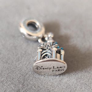 May include: Silver charm featuring a detailed replica of the Sleeping Beauty Castle at Disneyland Paris. The charm has blue enamel accents and the inscription "Disneyland Paris" on the bottom.