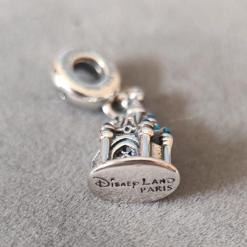 May include: Silver charm featuring a detailed replica of the Sleeping Beauty Castle at Disneyland Paris. The charm has blue enamel accents and the inscription "Disneyland Paris" on the bottom.