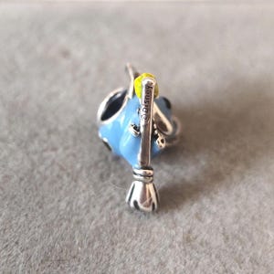 May include: A blue and silver charm with a yellow accent. The charm features a stylized design with a broom and the text "&copy;Disney". The charm is likely made of metal and enamel.