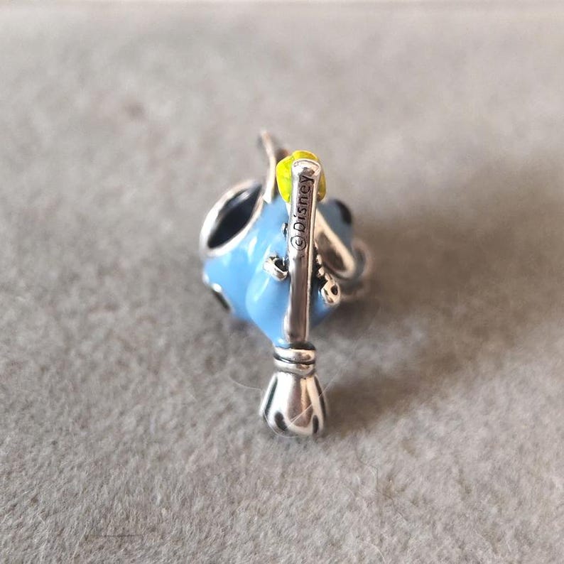 May include: A blue and silver charm with a yellow accent. The charm features a stylized design with a broom and the text "&copy;Disney". The charm is likely made of metal and enamel.
