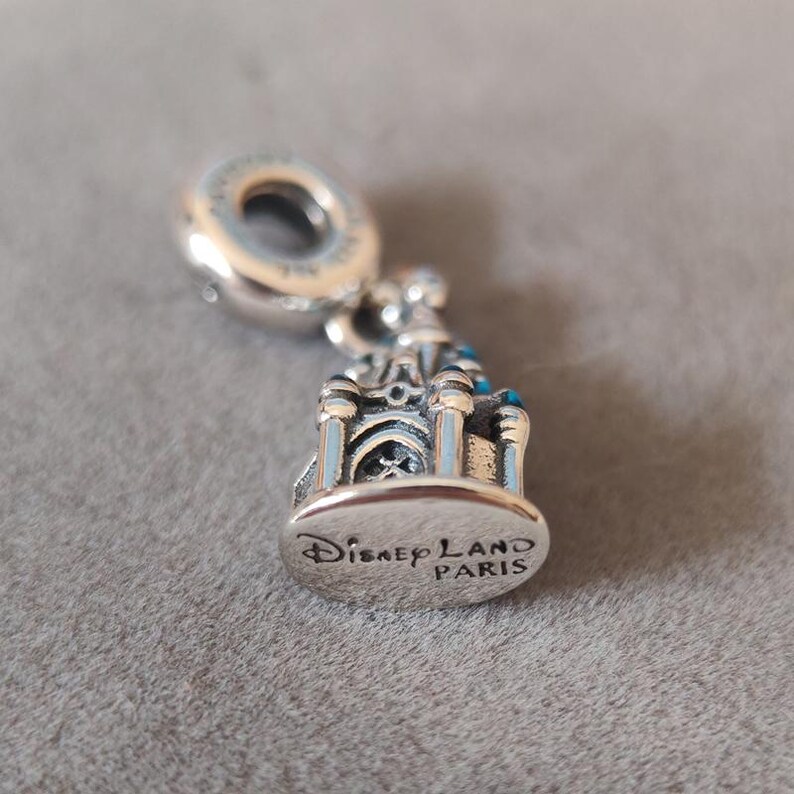 May include: Silver charm featuring a detailed replica of the Sleeping Beauty Castle at Disneyland Paris. The charm has a blue and white color scheme and the words "Disneyland Paris" are engraved on the bottom.