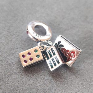 Puede incluir: Un charm de plata con un anillo con gemas rojas, un bloque rectangular con gemas rojas y verdes, y un peque&ntilde;o libro con un dise&ntilde;o floral rojo. Marcado con 'S 925'.