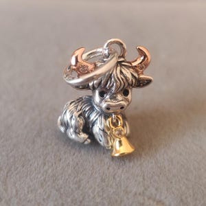 Puede incluir: Un charm plateado con forma de vaca de las Highlands. El charm tiene cuernos de color oro rosa y una campana dorada con una piedra transparente. La vaca tiene pelo detallado y un lazo para sujetarlo a una pulsera o collar.