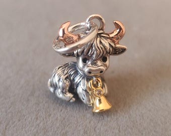 May include: A silver-colored charm in the shape of a Highland cow. The charm has rose gold-colored horns and a gold-colored bell with a clear stone. The cow has detailed fur and a loop for attaching to a bracelet or necklace.