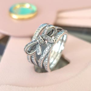 Puede incluir: Un anillo de plata con un diseño de lazo brillante. El anillo está hecho de múltiples bandas que están entrelazadas y engastadas con piedras de circonita cúbica transparentes.