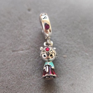Könnte beinhalten: Ein silberner Charm mit einem koreanischen Puppen-Design. Die Puppe trägt ein türkisfarbenes Oberteil und einen roten Rock. Der Charm hat eine Schlaufe mit den Buchstaben "KO" und einem roten Edelstein. Die Puppe hat einen roten Edelstein auf dem Kopf.