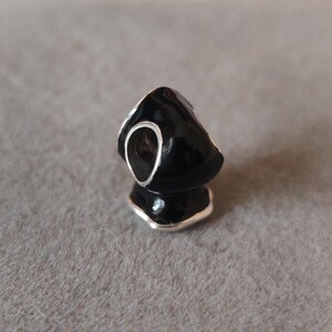 May include: A black charm with a silver-colored trim and a large, open oval shape. The charm has a glossy finish and is reflected in the surface below. The charm is likely a jewelry component, such as a bead or pendant.