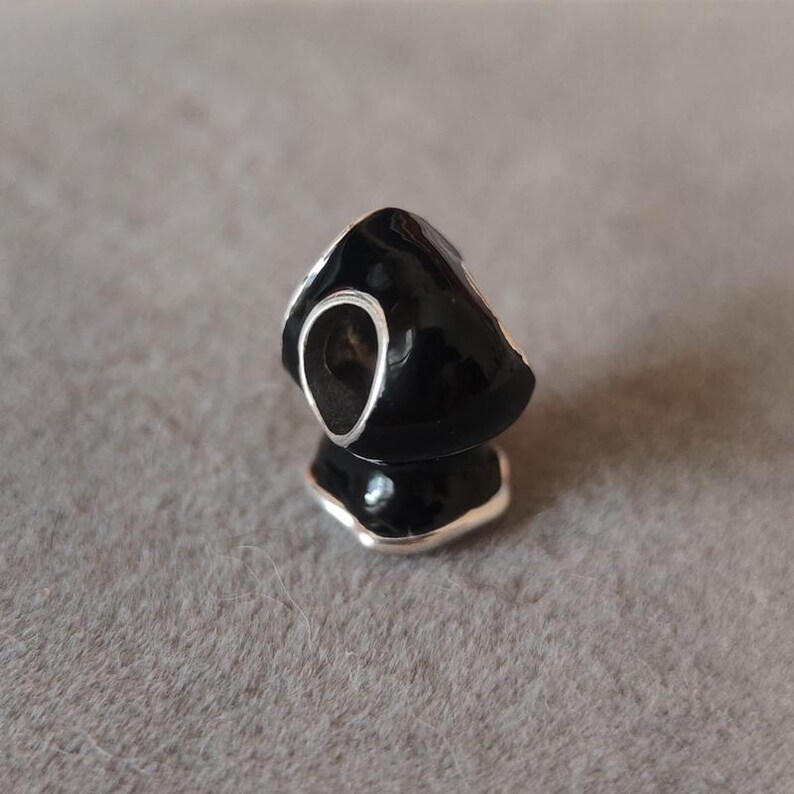 May include: A black charm with a silver-colored trim and a large, open oval shape. The charm has a glossy finish and is reflected in the surface below. The charm is likely a jewelry component, such as a bead or pendant.