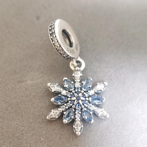 Op de afbeelding: Een zilveren bedel met een blauw en wit sneeuwvlokontwerp. De bedel heeft een kleine lus aan de bovenkant om aan een armband of ketting te bevestigen. De sneeuwvlok is versierd met heldere en blauwe kristallen.