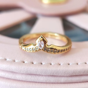 May include: A gold-colored ring with a crown-like design. The ring features a central, oval-shaped, clear gemstone and is encrusted with smaller, clear gemstones along the band. The ring is set on a pink surface.