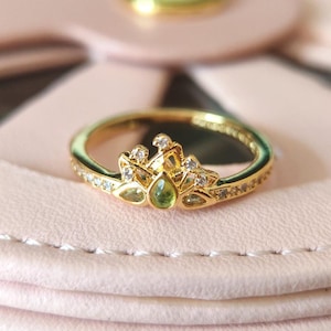 Op de afbeelding: Een goudkleurige ring met een kroonachtig ontwerp, met een peervormige groene edelsteen in het midden. De ring is versierd met kleine, heldere edelstenen en ligt op een roze ondergrond. De ringband is ook versierd met kleine edelstenen.