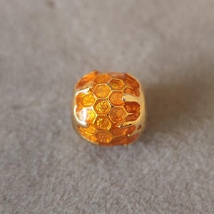 May include: A gold-coloured bead featuring a honeycomb pattern. The bead is spherical, with a series of hexagonal cells in a warm orange shade. The bead is likely metal and has a glossy finish.