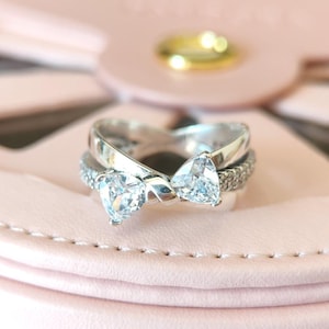 Könnte beinhalten: Ein silberner Ring mit zwei herzförmigen, klaren Steinen, die in einem verdrehten Band gefasst sind. Der Ring liegt auf einem rosa Hintergrund.