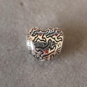 May include: A silver heart-shaped charm with a black, abstract design and small red accents. The charm has the words "With Heart" engraved on the side. The charm is on a gray background.