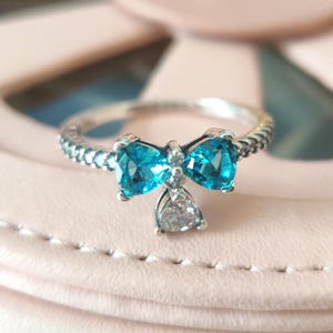 May include: A silver ring with a bow design. The bow features two heart-shaped, light blue gemstones and a pear-shaped, clear gemstone. The ring band has a textured, beaded pattern. The ring is a piece of jewellery.