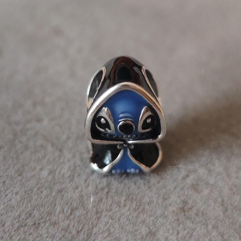 May include: A silver charm featuring a blue and black character design. The charm has a detailed face with large eyes and a small nose. The character's head is blue, and the charm has a shiny silver finish.