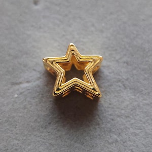 Puede incluir: Encanto de estrella de metal dorado con un diseño en capas. La estrella tiene un centro hueco y está hecha de múltiples capas de metal.