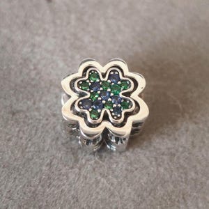 Könnte beinhalten: Ein silberner Charm mit einem vierblättrigen Kleeblatt-Design. Das Kleeblatt ist mit grünen und blauen Edelsteinen gefüllt. Der Charm hat ein detailliertes, geschichtetes Design und ist vor grauem Hintergrund platziert.