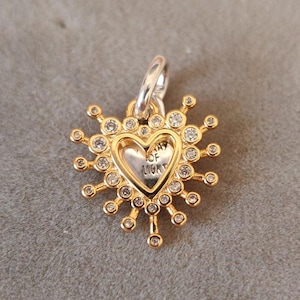Puede incluir: Colgante dorado con un centro en forma de corazón, rodeado de cristales brillantes y radios. El corazón presenta la inscripción "Ray of Light". El colgante tiene una fianza plateada para sujetarlo a una cadena o pulsera.