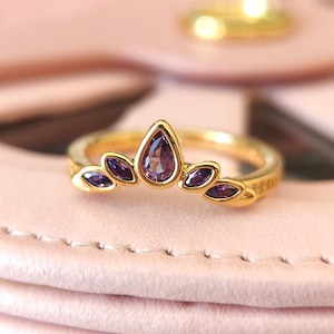 May include: A gold-colored ring with a teardrop-shaped purple gemstone at the center, surrounded by smaller purple gemstones in a leaf-like design. The ring is set against a soft pink background, showcasing its intricate details and craftsmanship.