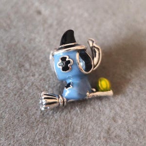 May include: A blue charm with a witch hat and broom. The charm features a cartoon character with a black witch hat, blue body, and silver broom. A small yellow ball is attached to the broom. The charm is likely made of metal and enamel.