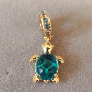 Könnte beinhalten: Goldfarbener Anhänger in Form einer Meeresschildkröte. Die Schildkröte hat einen türkisblauen Panzer mit grünem Muster und goldenen Details. Die Schlaufe des Anhängers ist mit blauen und türkisfarbenen Edelsteinen verziert.
