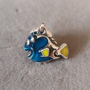 May include: A silver charm in the shape of a blue and yellow fish. The fish has a silver eye and a yellow fin.