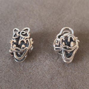 K&ouml;nnte beinhalten: Zwei silberfarbene Charms, die jeweils ein Cartoon-Entengesicht darstellen. Ein Charm hat eine Schleife auf dem Kopf, der andere einen Hut. Beide Charms haben einen l&auml;chelnden Ausdruck und sind mit dunklen Akzenten versehen.