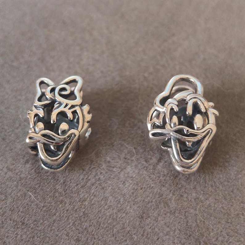 K&ouml;nnte beinhalten: Zwei silberfarbene Charms, die jeweils ein Cartoon-Entengesicht darstellen. Ein Charm hat eine Schleife auf dem Kopf, der andere einen Hut. Beide Charms haben einen l&auml;chelnden Ausdruck und sind mit dunklen Akzenten versehen.