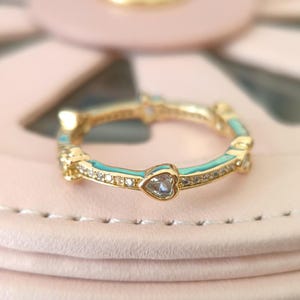 Puede incluir: Un anillo de oro con una banda de esmalte turquesa y peque&ntilde;os acentos en forma de coraz&oacute;n con piedras transparentes. El anillo est&aacute; sobre una superficie rosa.