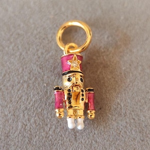 May include: A gold-colored charm in the shape of a nutcracker soldier. The charm has a pink hat with a star, black accents, and white boots. The charm is attached to a gold-colored ring.