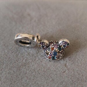 May include: A silver charm in the shape of Mickey Mouse's head, encrusted with a variety of colorful gemstones. The charm has a loop for attaching to a bracelet or necklace. The charm is on a gray surface.
