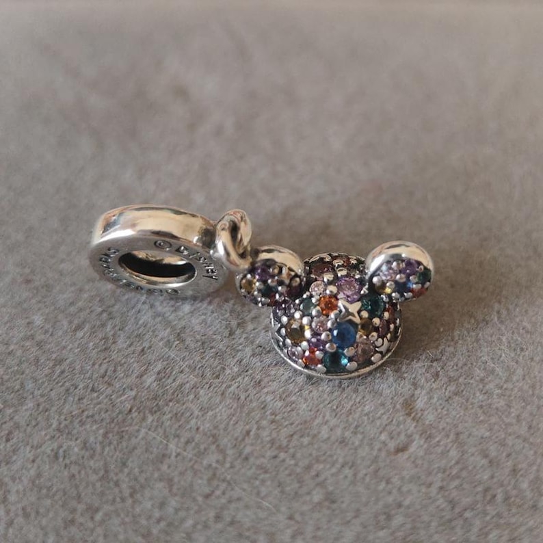 May include: A silver charm in the shape of Mickey Mouse's head, encrusted with a variety of colorful gemstones. The charm has a loop for attaching to a bracelet or necklace. The charm is on a gray surface.