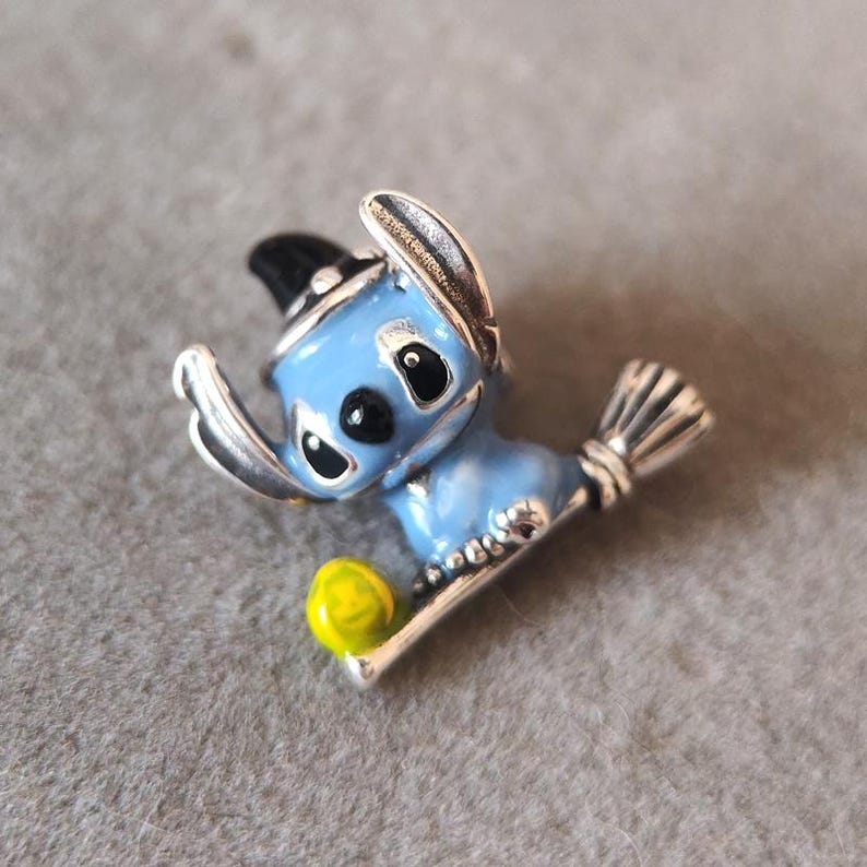 May include: A blue enamel and silver-tone charm featuring a cartoon character wearing a black witch hat and holding a broom. The charm has a yellow ball and is set against a gray background. The charm is likely for a bracelet or necklace.