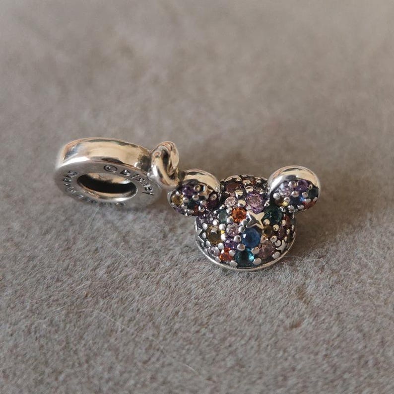 May include: A silver charm in the shape of Mickey Mouse's head, encrusted with colorful gemstones. The charm has a loop for attaching to a bracelet or necklace. The charm is on a gray surface.