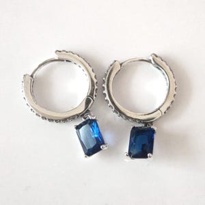 Peut inclure: Une paire de boucles d'oreilles créoles en argent avec une pierre précieuse bleue suspendue. Les boucles d'oreilles sont simples et élégantes.
