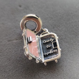 Puede incluir: Un charm de plata con forma de maleta abierta con las palabras "Ready to Explore" grabadas en la parte delantera. El interior es rosa y el charm tiene un lazo para sujetarlo a una pulsera o collar.